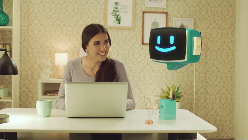 Mulher sentada na frente de um notebook e uma cabeça de robô sorridente suspensa no ar ao lado da mulher.