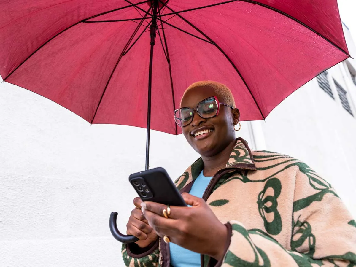 Wanita memegang payung merah, berjalan di luar dengan tersenyum sambil melihat teleponnya