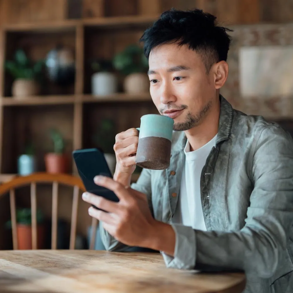 Um homem sentado à mesa bebendo uma caneca de café enquanto usa o celular