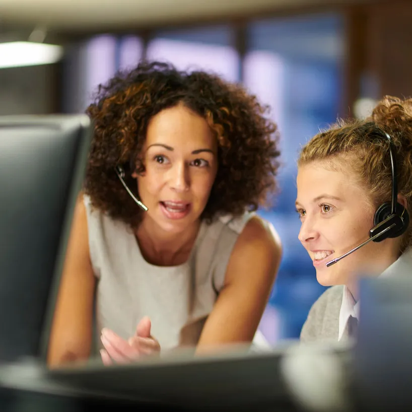 Duas mulheres usando headsets olhando para o monitor de um computador em frente a elas.